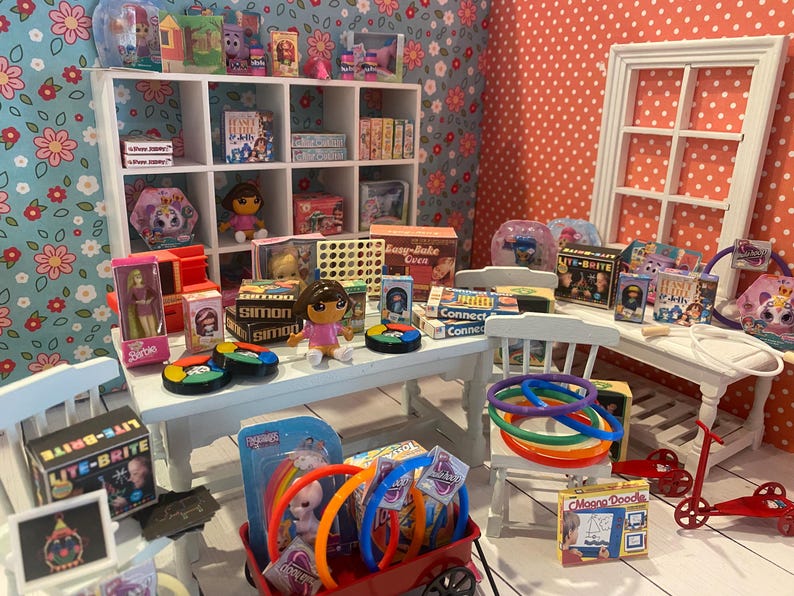 Op de afbeelding: Een miniatuur speelgoedwinkeldisplay met diverse speelgoed en spelletjes. De display bevat poppen, bordspellen en een Lite-Brite set. Het kleurenschema is helder en speels, met een rode wagen vol kleurrijke ringen en een rode scooter.