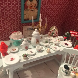 May include: Miniature Christmas-themed tea set with a white table, featuring a cake, teapot, cups, and plates. The set includes a Santa Claus picture, a gingerbread house, and a "Merry Christmas" sign. The background has a red floral wallpaper.