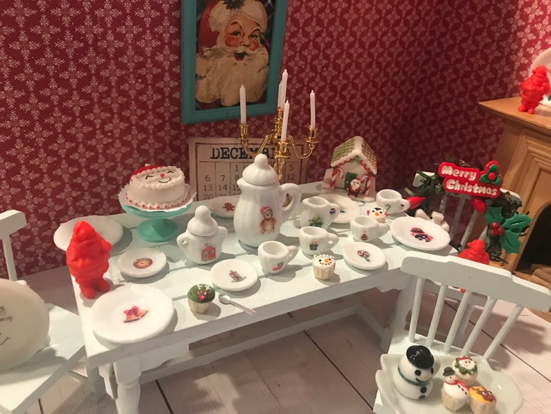 May include: Miniature Christmas-themed tea set with a white table, featuring a cake, teapot, cups, and plates. The set includes a Santa Claus picture, a gingerbread house, and a "Merry Christmas" sign. The background has a red floral wallpaper.