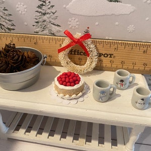 May include: A white miniature table with a red and white cake, a white wreath with a red bow, a silver bucket with pine cones, and three white mugs with snowman designs. The table has a lower shelf with white slats.