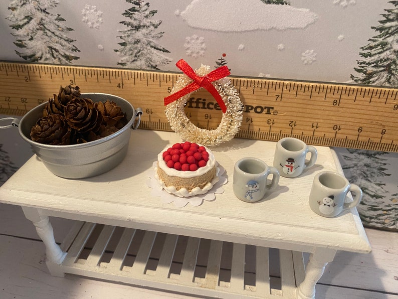 May include: A white miniature table with a red and white cake, a white wreath with a red bow, a silver bucket with pine cones, and three white mugs with snowman designs. The table has a lower shelf with white slats.