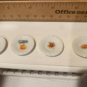 May include: Four small, white decorative plates with Thanksgiving-themed designs. One plate features a pilgrim hat, another has a pumpkin and the word "Thanksgiving", a third has a maple leaf, and the last has a roasted turkey.