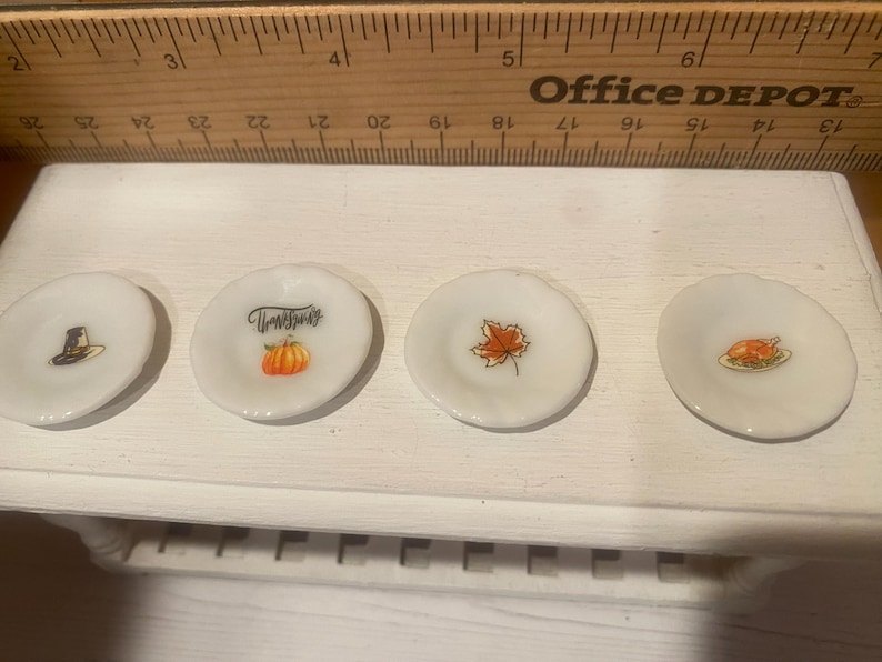 May include: Four small, white decorative plates with Thanksgiving-themed designs. One plate features a pilgrim hat, another has a pumpkin and the word "Thanksgiving", a third has a maple leaf, and the last has a roasted turkey.