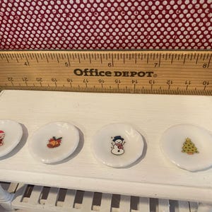 May include: Four small, white, decorative plates with Christmas-themed designs. Each plate features a different image: Santa Claus, a bell, a snowman, and a Christmas tree. The plates are arranged on a white surface.