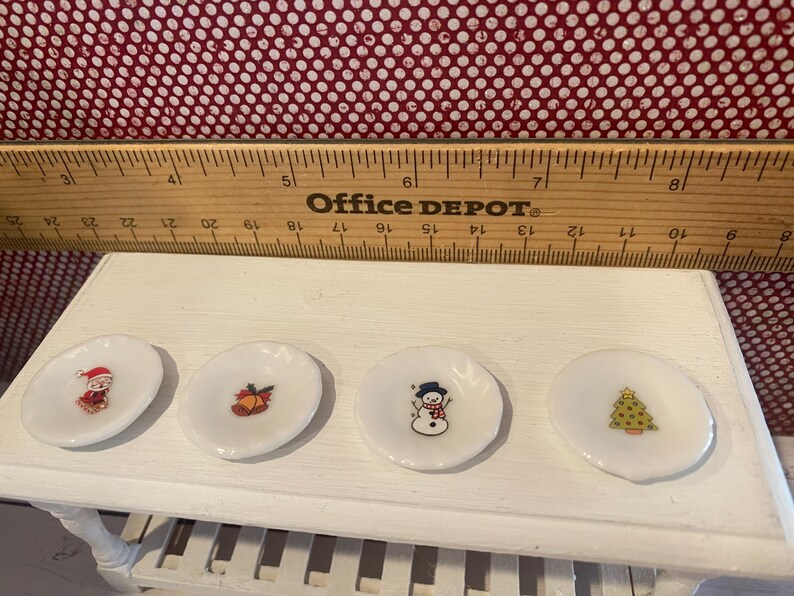 May include: Four small, white, decorative plates with Christmas-themed designs. Each plate features a different image: Santa Claus, a bell, a snowman, and a Christmas tree. The plates are arranged on a white surface.