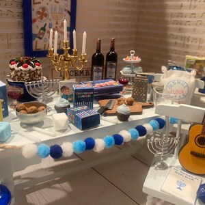 May include: A miniature table setting for a Hanukkah celebration with blue and white decorations, a menorah, a chocolate cake, and a guitar. The table is decorated with a string of blue and white pom-poms. The table also includes a box of Chanukah candles, a small book, and a bag of flour.