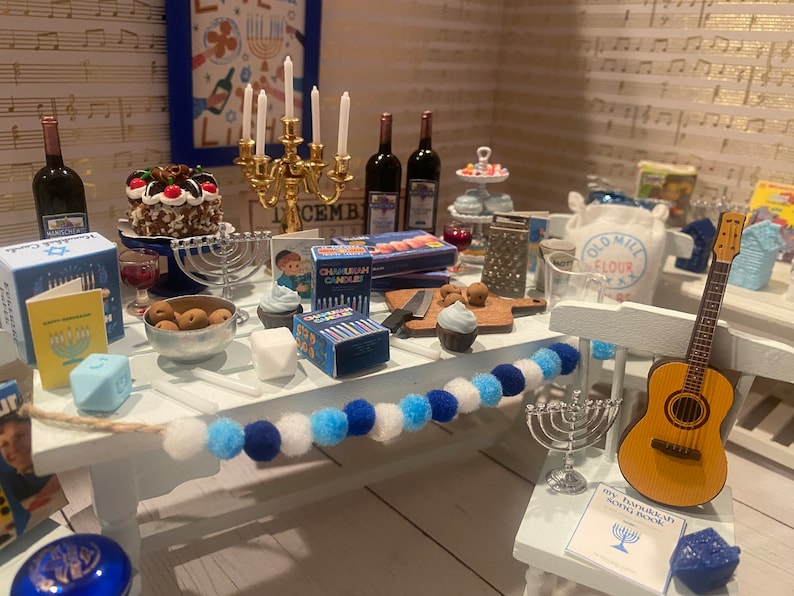 May include: A miniature table setting for a Hanukkah celebration with blue and white decorations, a menorah, a chocolate cake, and a guitar. The table is decorated with a string of blue and white pom-poms. The table also includes a box of Chanukah candles, a small book, and a bag of flour.