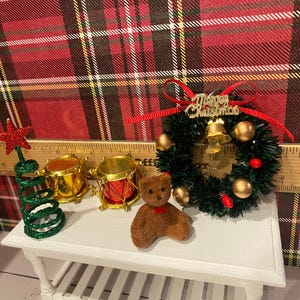 Puede incluir: Decoraciones navide&ntilde;as en miniatura sobre una mesa blanca. Incluye un &aacute;rbol de estrella verde y roja, tambores dorados, un oso de peluche marr&oacute;n con un lazo rojo y una corona navide&ntilde;a con una campana y adornos dorados.