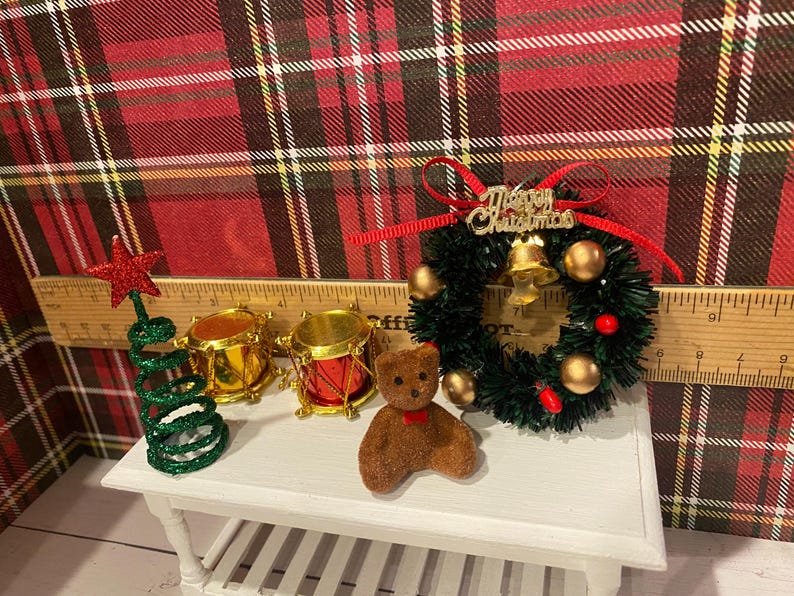 Puede incluir: Decoraciones navide&ntilde;as en miniatura sobre una mesa blanca. Incluye un &aacute;rbol de estrella verde y roja, tambores dorados, un oso de peluche marr&oacute;n con un lazo rojo y una corona navide&ntilde;a con una campana y adornos dorados.