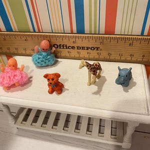 May include: A collection of miniature toys, including two baby dolls in pink and blue crocheted outfits, a small orange teddy bear, a giraffe, and a blue elephant. The toys are arranged on a white table, with a wooden ruler in the background.