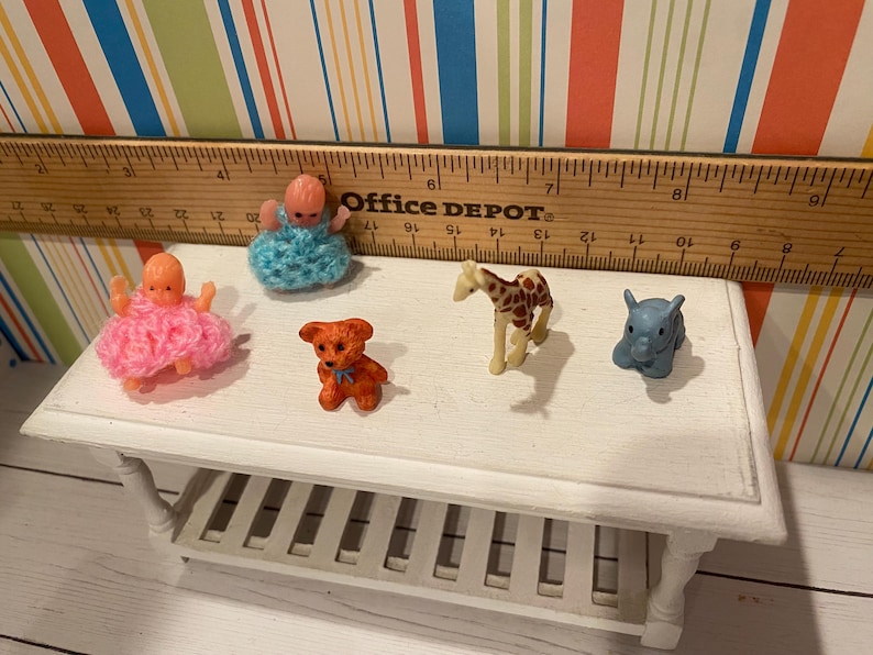 May include: A collection of miniature toys, including two baby dolls in pink and blue crocheted outfits, a small orange teddy bear, a giraffe, and a blue elephant. The toys are arranged on a white table, with a wooden ruler in the background.