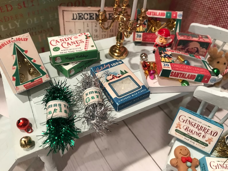 May include: A miniature Christmas scene with various holiday decorations. Boxes of candy canes, Christmas lights, and gingerbread baking are displayed on a white table. Green and silver tinsel garland and a candle holder add to the festive atmosphere.