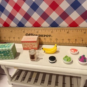 May include: Miniature food items on a white table, including ice cream boxes, a mug of beer, a banana, an Oreo, and small dishes of fruit. A ruler and a red, white, and blue checkered background complete the scene. The ice cream boxes read "Sclater's Ice Cream" and "Home Maid Ice Cream."