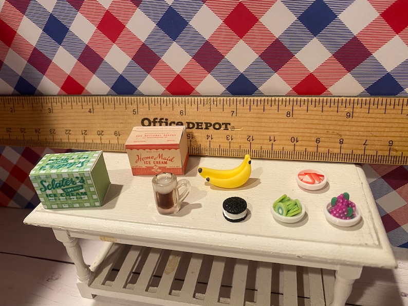 May include: Miniature food items on a white table, including ice cream boxes, a mug of beer, a banana, an Oreo, and small dishes of fruit. A ruler and a red, white, and blue checkered background complete the scene. The ice cream boxes read "Sclater's Ice Cream" and "Home Maid Ice Cream."