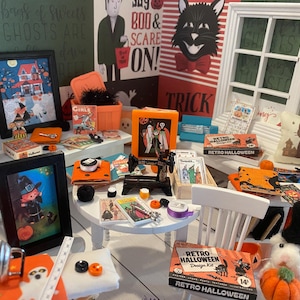 Puede incluir: Una exhibición en miniatura con temática de Halloween con arte enmarcado, cajas decorativas y una pequeña máquina de coser. La paleta de colores incluye naranja, negro y blanco, con una caja de kit de diseño retro y un cojín con una silueta de gato.