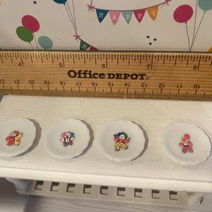 May include: Four small, white, scalloped-edge plates, each with a different clown illustration. A wooden ruler rests above the plates. The background features a decorative banner with balloons and the word "HAPPY".