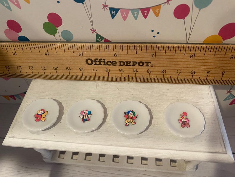 May include: Four small, white, scalloped-edge plates, each with a different clown illustration. A wooden ruler rests above the plates. The background features a decorative banner with balloons and the word "HAPPY".