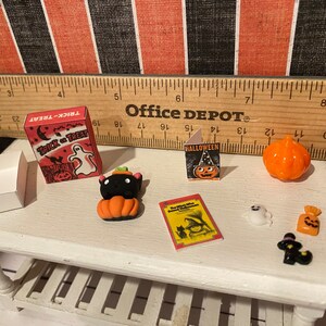 May include: A white table with a variety of Halloween themed miniature items, including a box of trick or treat candy, a black cat with a pumpkin, a small book with the word "Halloween" on the cover, a small orange pumpkin, a ghost, a bag of candy, and a black cat with a witch's hat.