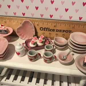 May include: A miniature ceramic tea set and dessert collection featuring pink heart-shaped plates, small cups with red heart patterns, a white sugar bowl, a rectangular tray with rose-topped cupcakes, and two slices of chocolate cake with cherries.