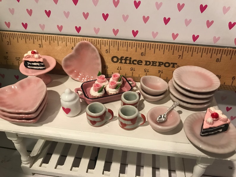 May include: A miniature ceramic tea set and dessert collection featuring pink heart-shaped plates, small cups with red heart patterns, a white sugar bowl, a rectangular tray with rose-topped cupcakes, and two slices of chocolate cake with cherries.