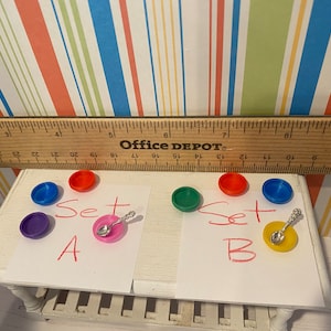 May include: A miniature set of colorful plastic dishes, including red, blue, green, and purple, with a small spoon on a pink plate. The text "Set A" and "Set B" are written on paper.