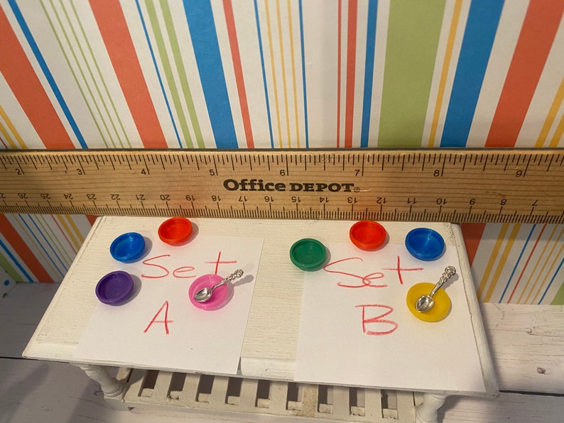 May include: A miniature set of colorful plastic dishes, including red, blue, green, and purple, with a small spoon on a pink plate. The text "Set A" and "Set B" are written on paper.