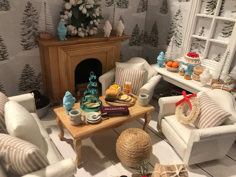 May include: A miniature living room scene with a white fireplace, two white armchairs with striped cushions, a coffee table with a game of Pictionary, a blue Christmas tree, and a blue snowman figurine.