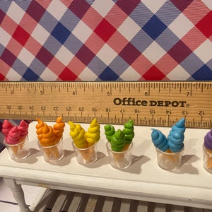 May include: Miniature ice cream cones in various colors, including pink, orange, yellow, green, blue, and purple, displayed in clear plastic cups. The cones sit on a white table, with a ruler and a red, white, and blue checkered background.