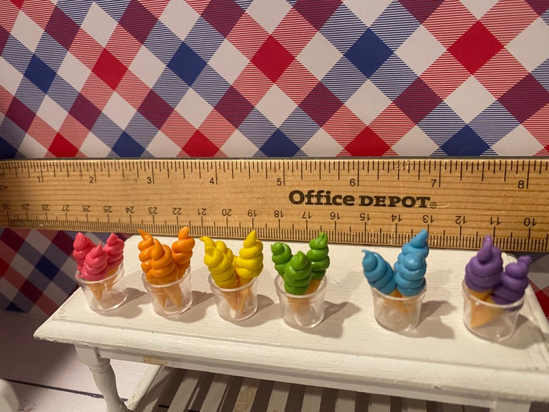 May include: Miniature ice cream cones in various colors, including pink, orange, yellow, green, blue, and purple, displayed in clear plastic cups. The cones sit on a white table, with a ruler and a red, white, and blue checkered background.