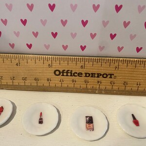 May include: Four small white ceramic plates, each featuring a different miniature illustration of beauty products including a perfume bottle, lipstick, compact, and nail polish, positioned below a wooden ruler measuring approximately 8 inches.