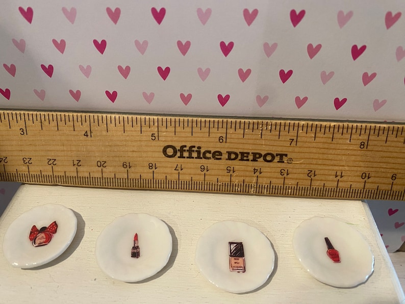 May include: Four small white ceramic plates, each featuring a different miniature illustration of beauty products including a perfume bottle, lipstick, compact, and nail polish, positioned below a wooden ruler measuring approximately 8 inches.