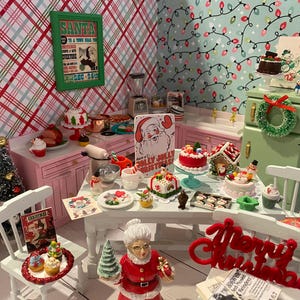 May include: A miniature dollhouse kitchen decorated for Christmas. The kitchen features a pink counter with a white countertop, a green cabinet with a wreath, and a white table with a red "Merry Christmas" sign. The table is set with miniature food and decorations, including a gingerbread house, a Santa Claus figurine, and a Christmas tree.