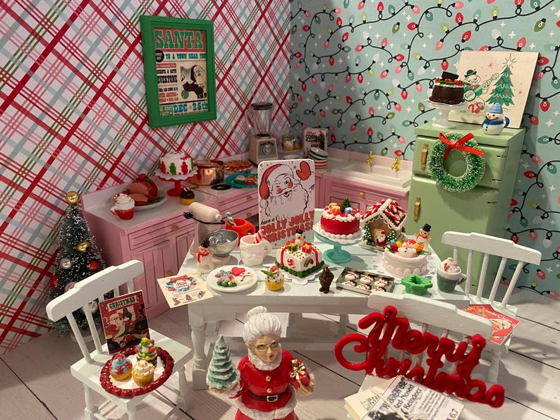 May include: A miniature dollhouse kitchen decorated for Christmas. The kitchen features a pink counter with a white countertop, a green cabinet with a wreath, and a white table with a red "Merry Christmas" sign. The table is set with miniature food and decorations, including a gingerbread house, a Santa Claus figurine, and a Christmas tree.