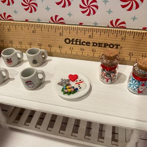 Pode incluir: Uma mesa branca com quatro canecas brancas em miniatura com desenhos de Natal, um prato pequeno com um boneco de neve e uma &aacute;rvore de Natal, e dois pequenos frascos de vidro com bast&otilde;es de caramelo vermelhos e brancos.