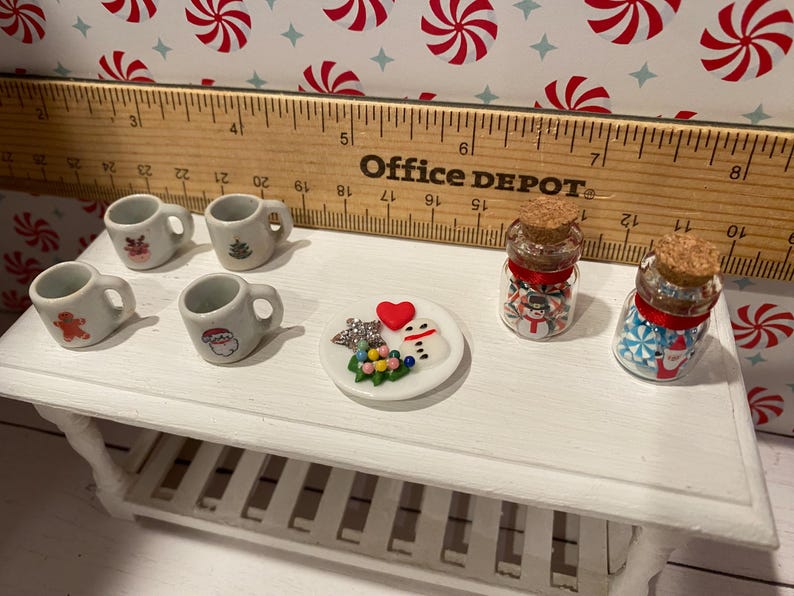 Pode incluir: Uma mesa branca com quatro canecas brancas em miniatura com desenhos de Natal, um prato pequeno com um boneco de neve e uma &aacute;rvore de Natal, e dois pequenos frascos de vidro com bast&otilde;es de caramelo vermelhos e brancos.
