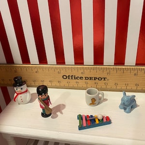 May include: A collection of miniature toys including a snowman, a soldier, a mug, a blue elephant, and a xylophone. The toys are arranged on a white surface with a ruler and a red and white striped background.