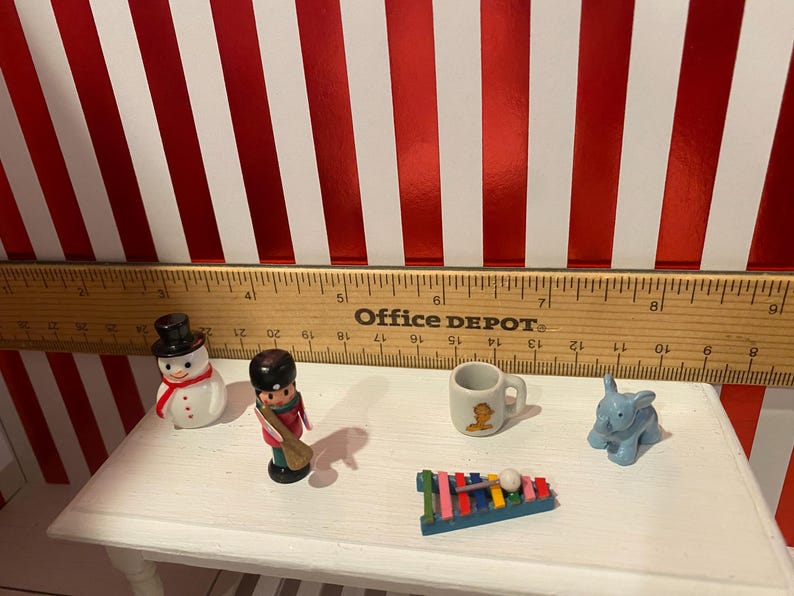 May include: A collection of miniature toys including a snowman, a soldier, a mug, a blue elephant, and a xylophone. The toys are arranged on a white surface with a ruler and a red and white striped background.