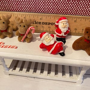 May include: A miniature white table displays Christmas-themed figurines. Two brown reindeer with red bows, a candy cane, two Santa Claus figures in red suits, and a brown teddy bear with a red bow are arranged on the table.