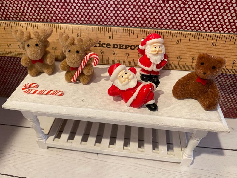 May include: A miniature white table displays Christmas-themed figurines. Two brown reindeer with red bows, a candy cane, two Santa Claus figures in red suits, and a brown teddy bear with a red bow are arranged on the table.