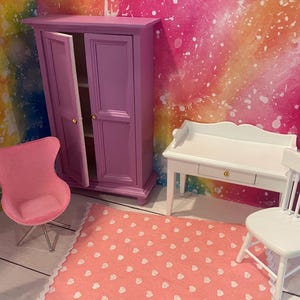 K&ouml;nnte beinhalten: Ein Puppenhaus-Schlafzimmerset mit einem rosa Kleiderschrank, einem wei&szlig;en Schreibtisch mit Schublade und einem rosa eif&ouml;rmigen Stuhl. Das Set befindet sich auf einem rosa-wei&szlig;en Teppich.