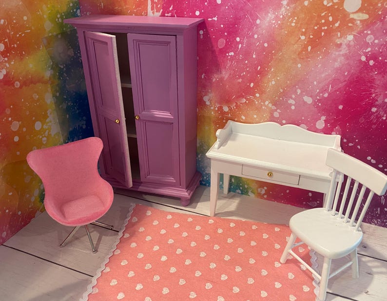 K&ouml;nnte beinhalten: Ein Puppenhaus-Schlafzimmerset mit einem rosa Kleiderschrank, einem wei&szlig;en Schreibtisch mit Schublade und einem rosa eif&ouml;rmigen Stuhl. Das Set befindet sich auf einem rosa-wei&szlig;en Teppich.