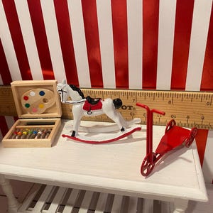 May include: Miniature toys on a white table. Includes a white rocking horse with a red saddle, a red scooter, and a wooden art set with a paint palette and colored pencils. The background is red and white striped.