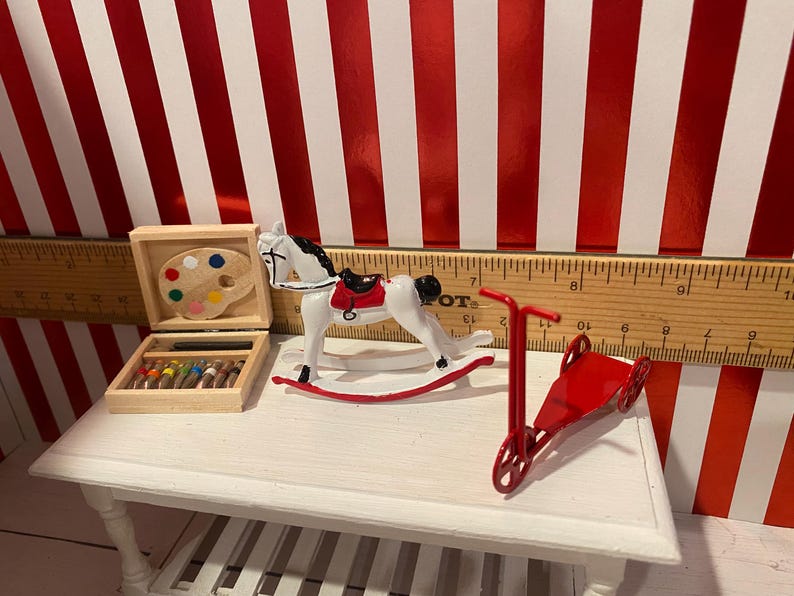 May include: Miniature toys on a white table. Includes a white rocking horse with a red saddle, a red scooter, and a wooden art set with a paint palette and colored pencils. The background is red and white striped.