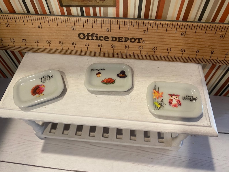 May include: Three small, rectangular ceramic dishes with fall-themed designs. One dish features a turkey, another a pumpkin and hat, and the third an owl with leaves. The dishes are on a white shelf with a wooden ruler.