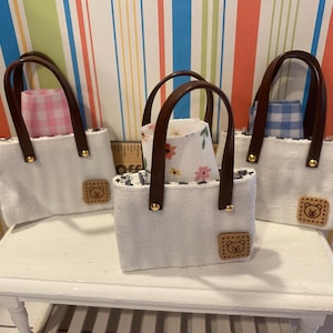 May include: Three miniature white tote bags with brown handles and bear-shaped patches. Each bag contains a patterned fabric lining, including pink gingham, blue gingham, and floral designs. The bags are displayed on a white surface.