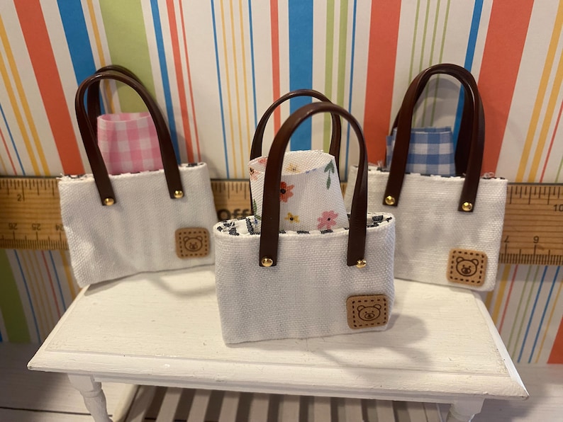 May include: Three miniature white tote bags with brown handles and bear-shaped patches. Each bag contains a patterned fabric lining, including pink gingham, blue gingham, and floral designs. The bags are displayed on a white surface.