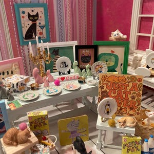 K&ouml;nnte beinhalten: Ein Miniatur-Puppenhauszimmer mit katzenthematischer Dekoration, darunter verschiedene kleine Katzenfiguren, gerahmte Katzenbilder, winzige Zierteller mit Katzenmotiven, ein goldener Kerzenst&auml;nder und Meow Mix-Schachteln auf einem wei&szlig;en Tisch.