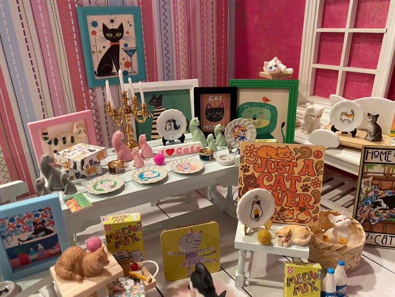 K&ouml;nnte beinhalten: Ein Miniatur-Puppenhauszimmer mit katzenthematischer Dekoration, darunter verschiedene kleine Katzenfiguren, gerahmte Katzenbilder, winzige Zierteller mit Katzenmotiven, ein goldener Kerzenst&auml;nder und Meow Mix-Schachteln auf einem wei&szlig;en Tisch.