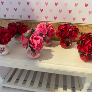 May include: Five miniature vases, each holding a cluster of artificial roses in shades of red or pink. The vases are white, pink, or red, and feature hand-painted heart motifs and the word love. A wooden ruler is visible in the background.