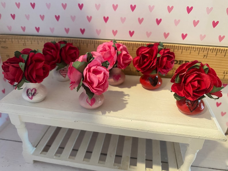 May include: Five miniature vases, each holding a cluster of artificial roses in shades of red or pink. The vases are white, pink, or red, and feature hand-painted heart motifs and the word love. A wooden ruler is visible in the background.